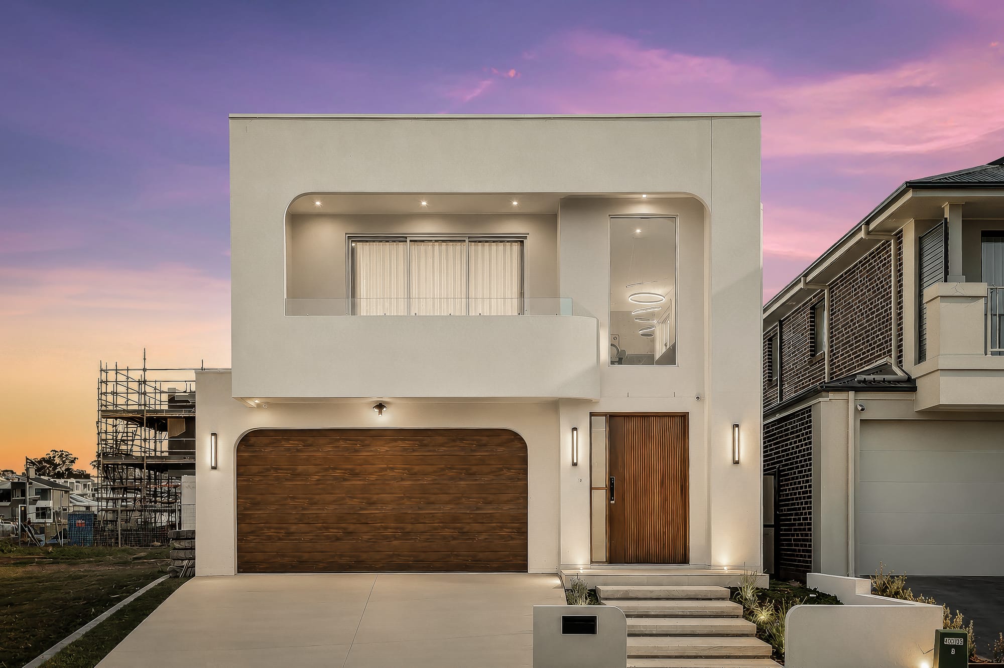 Double-storey home at Gelderland Drive, Box Hill at dusk, with a rendered façade, a timber-look garage door and a timber front door